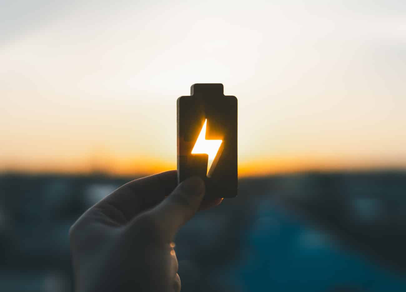Glowing battery symbol held up against a sunset, representing solar stored for the evening Glowing battery symbol held up against a sunset, representing solar stored for the evening
